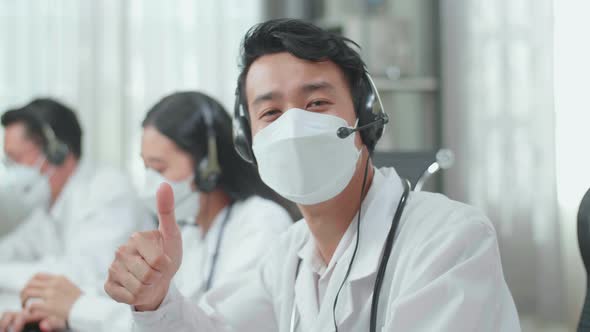 Asian Man Doctor Wearing Headsets And Masks Working As Call Centre Agent Look Up And Thumbs Up