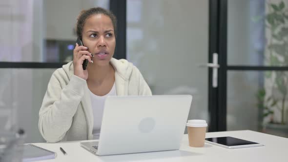 Angry African Woman Talking on Smartphone While Using Laptop in Office