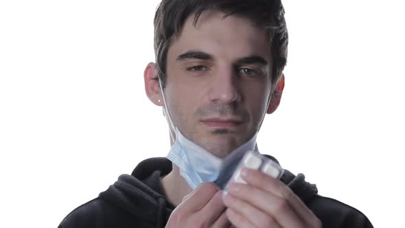 A Portrait of a Young Man in a Medical Mask Who Takes Off His Mask and Takes a Pill on a White