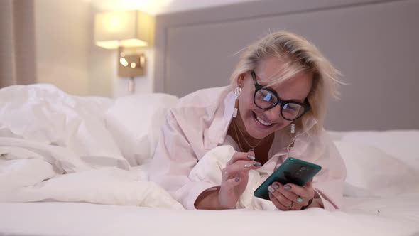 Cheerful Lady Is Typing Comments in Social Net Using Smartphone Lying in Bed at Evening Time