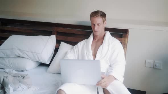 Handsome Man in White Bathrobe Lies on Bed in His Apartment and Uses Laptop for Work