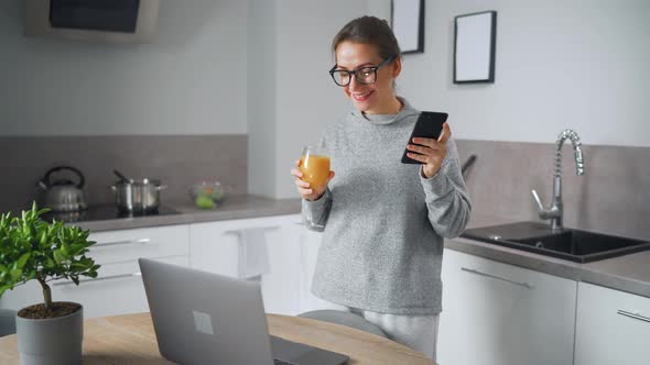 Woman with a Glass of Juice in Her Hand Stands in the Kitchen and Works at a Laptop Do Freelance