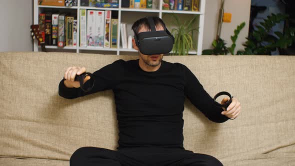 A man wearing VR glasses and black clothes is actively playing on the couch