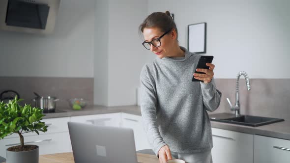 Woman with Smartphone in Hand Standing in Home Cozy Kitchen Use Laptop Do Freelance Work Looking