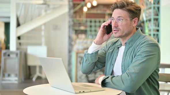 Middle Aged Man with Laptop Talking on Smartphone in Cafe