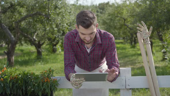 Young Farmer Using the Tablet Standing Behind the Fence in the Foreground, the Summer Garden