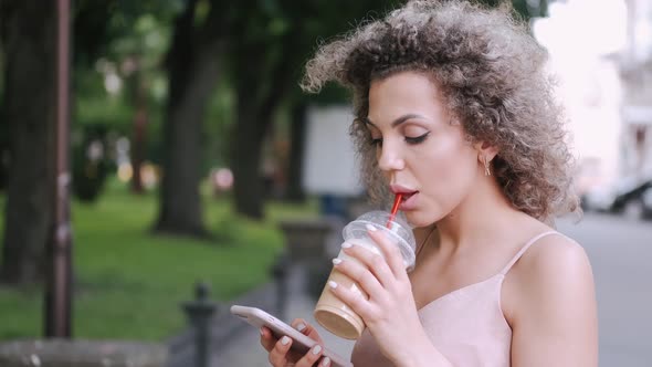 Attractive Young Woman Is Drinking a Cocktail and Using Smartphone in the Summer City
