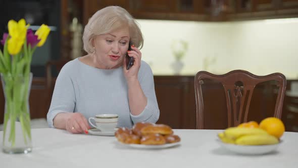 Carefree Smiling Beautiful Retiree Talking on Smartphone Drinking Herbal Tea in the Evening at Home