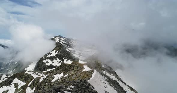 Forward Aerial Top View Over Cloudy Rocky Snowy Mountain in Sunny Day with Clouds