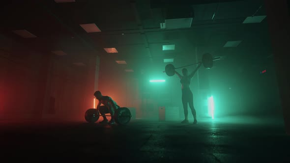 A Man and a Woman in a Colored Neon Red and Blue Light in Slow Motion Lift Heavy Barbells Over Their