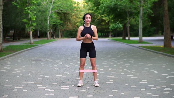 Young Woman Athlete Exersices with Resistance Band Doing Squats Outdoors at Local Green Public Park