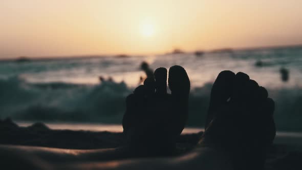 POV Silhouette of Feet of Young Man Lying on Sandy Beach By Ocean During Sunset