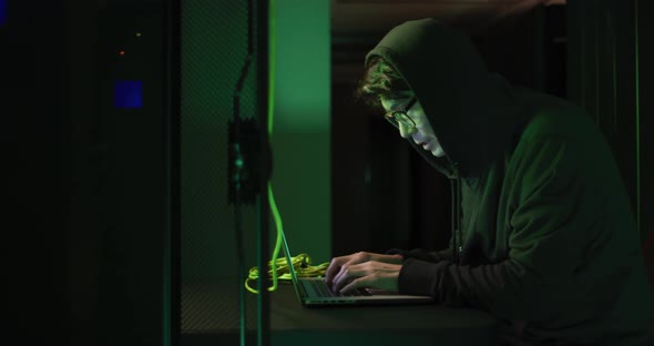 Asian male hacker in hoodie using laptop by computer servers
