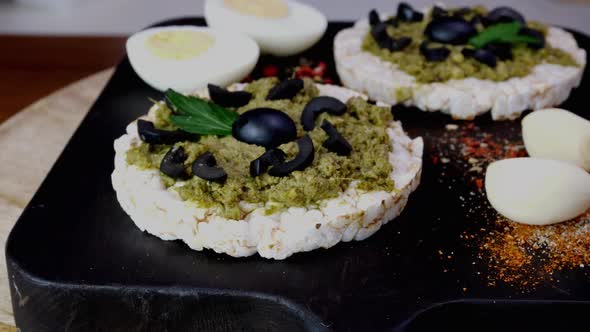 Rice cake toasts with spreading green pesto sauce and boiled eggs rotate slowly on cutting board.