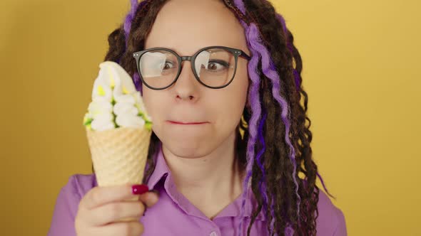 Young beautiful woman in glasses looking at ice cream, licking lips on yellow background.