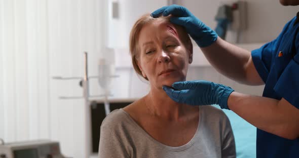 Male Doctor Examining Wound on Aged Woman Forehead in Clinic