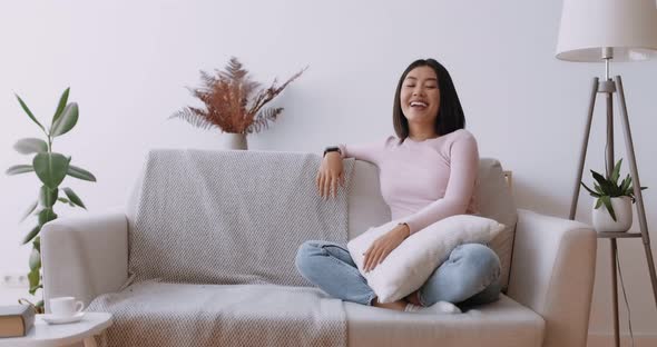 Domestic Comfort. Happy Asian Woman Sitting on Sofa at Home at Laughing at Camera