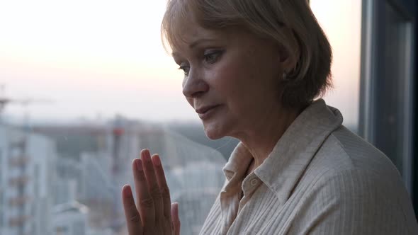 Mature Sad Woman Looks Out Window From Apartment Hoping For Miracle