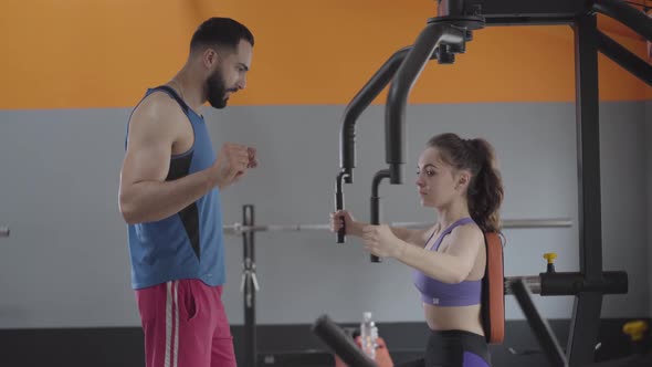 Serious Trainer Helping Young Slender Woman with Workout in Gym
