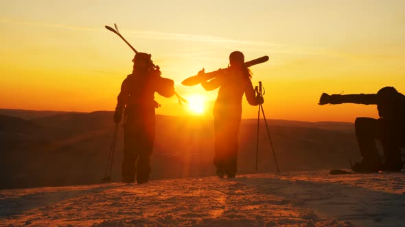 People with ski equipment walk along snowy hilltop at sunset