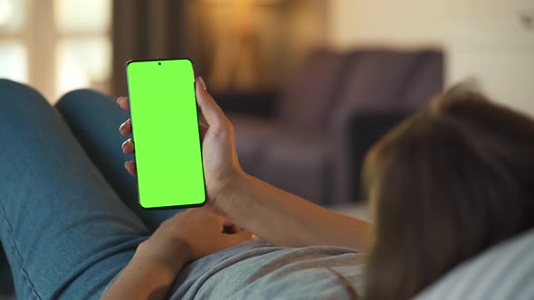 Woman at Home Lying on a Sofa and Using Smartphone with Green Mock-up Screen in Vertical Mode. Girl