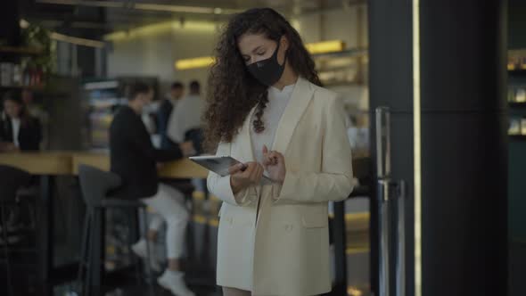 Busy Caucasian Businesswoman in Covid-19 Face Mask Using Tablet and Looking at Camera. Blurred