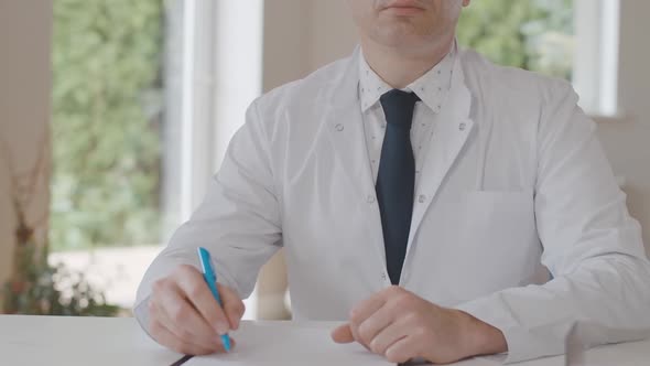 Unrecognizable Male Doctor Sitting at the Table with Documents. Confident Caucasian Physician Posing