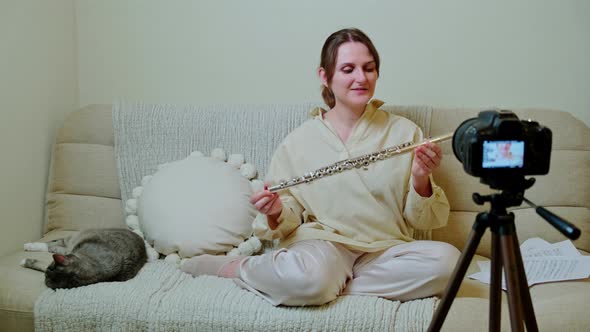 Happy woman musician with flute looking at video camera at home on sofa in living room