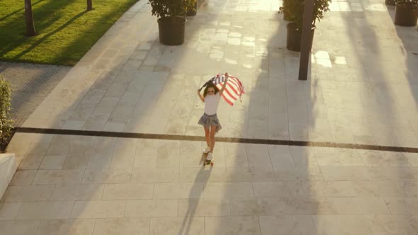 Young Active Caucasian Woman Rides Skateboard Holding American Flag Over Head