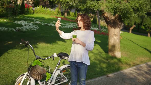 Portrait of a Young Woman Taking Selfie Photo with Cellphone Standing in the City Park Near Her City