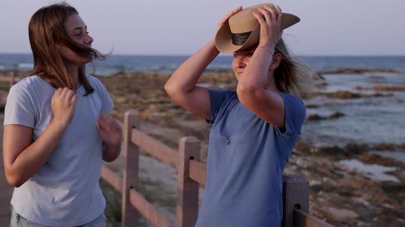 Woman standing on sidewalk by the sea at sunset near her teenage daughter and wearing straw hat