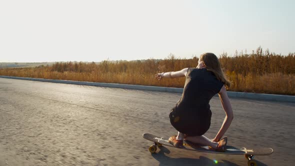 Squatting on a Longboard, Young Woman in a Dress Is Longboarding
