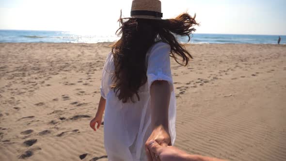 Follow Me Shot of Young Woman in Hat Pull Her Boyfriend on the Sea Shore. Girl Holding Male Hand and