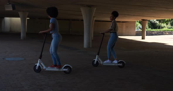 Two mixed race women riding electric scooter under bridge