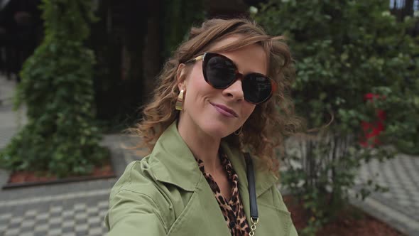 Young Woman with Long Curly Hair in Sunglasses Makes a Selfie on the Street