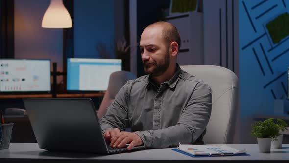 Overworked Businessman Typing Financial Statistics on Laptop Sitting at Desk Table