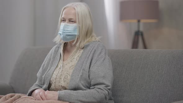 Medium Shot Portrait of Old Greyhaired Woman in Coronavirus Face Mask Sitting on Couch Looking at