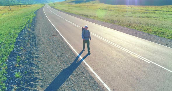 Flight Over Hitchhiker Tourist Walking on Asphalt Road. Huge Rural Valley at Summer Day. Backpack