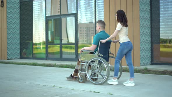 A Young Woman Is Carrying a Young Man in a Wheelchair. An Assistant Carries a Disabled Person in a