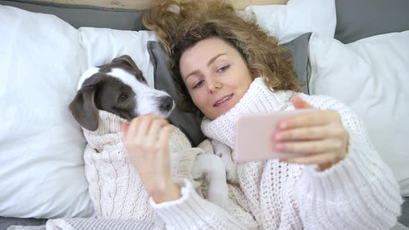 Young Woman Taking Selfie With Her Dog Using Smartphone Lying In Bed