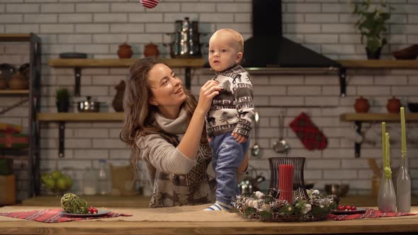 Pretty Young Woman Lifts Up the Baby in Her Arms Standing in Modern Kitchen Showing To Son Bright