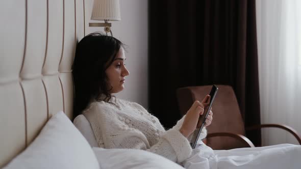 Young Beautiful Freelance Woman Sitting on Bed and Working on Tablet Screen Morning Time Working