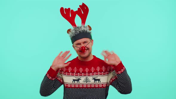 Positive Man in Christmas Sweater Deer Antlers Waves Hand Palm in Hello Gesture Welcomes Someone