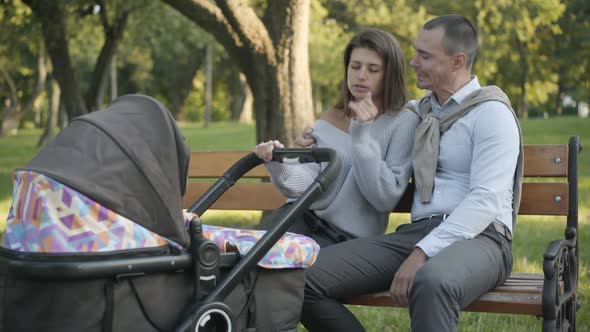 Exhausted Young Couple of Parents Sitting on Bench in Summer Park Rocking Baby Stroller, and Yawning