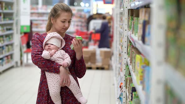 Woman Choosing Children Food with Little Baby Child Girl on Hands During Supermarket Shopping