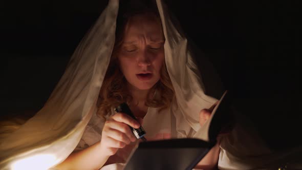 Woman Reads Book Under Blanket in the Dark and Illuminates the Pages of the Book with Flashlight