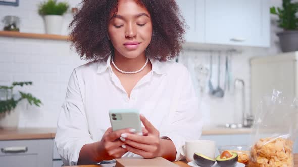 Young African American Woman Uses Phone Taking Healthy Breakfast Sits at Kitchen