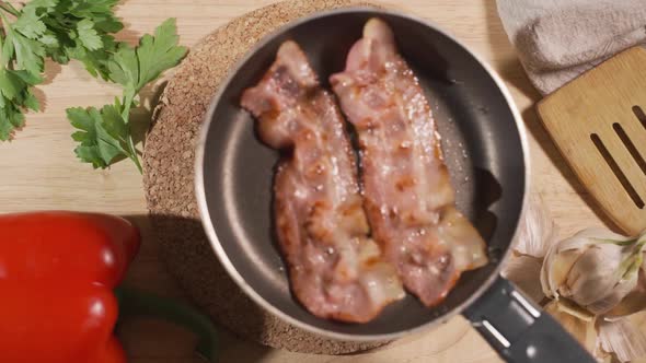 The Cook Puts on a Served Table a Frying Pan with Fried Eggs and Bacon. On the Table Are Greens