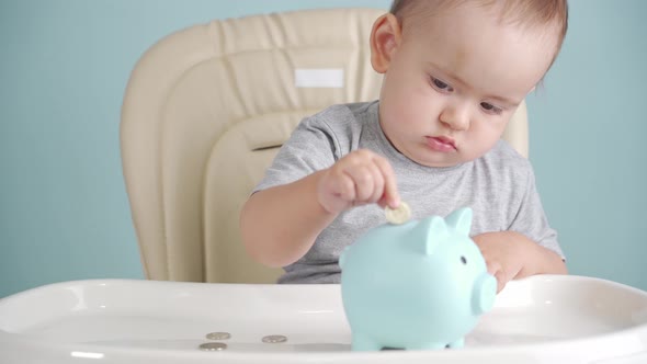 Toddler 12-17 months old throws a coin into a blue piggy bank sitting on a chair, selective focus