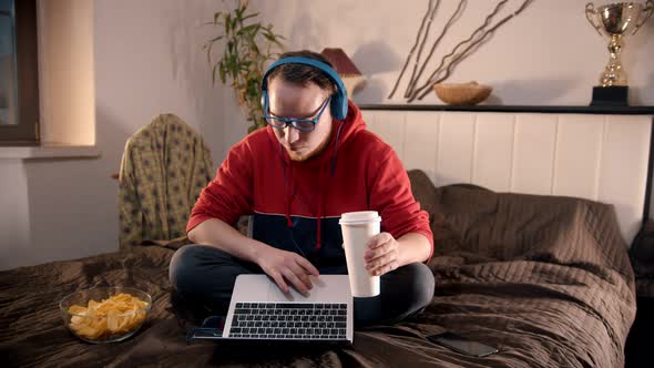 Young Man Sitting on the Bed Talking with Somebody Using Laptop and Drinking Coffee From the Paper